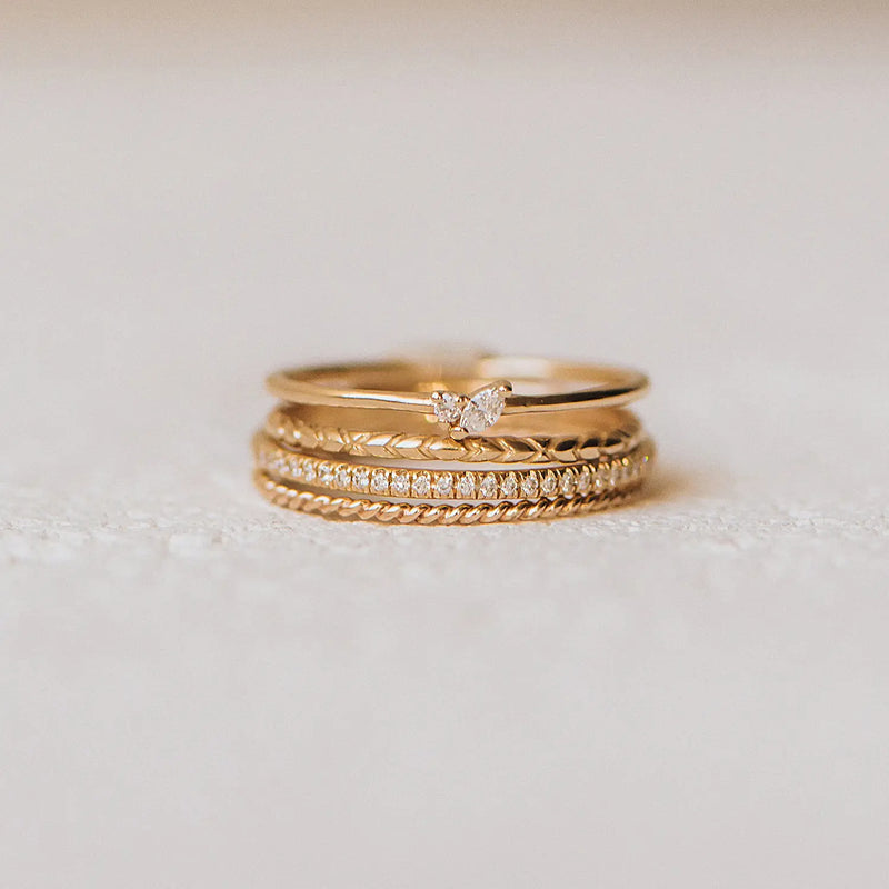 A stack of three bluboho twisted stacking rings - 10k yellow gold lies on a light, fabric surface. The bottom ring features a rope-like design, the middle ring is studded with small, round diamonds, and the top ring has a small, pear-shaped diamond solitaire set on it.