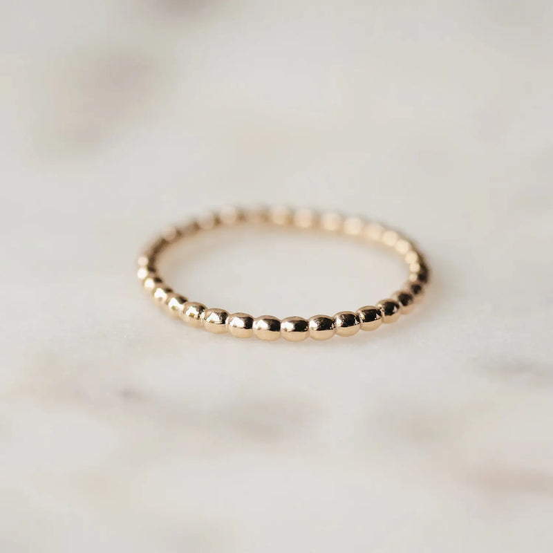 A close-up image of a delicate abacus ring - 10k yellow gold by bluboho, placed on a light-colored, slightly blurred marbled surface. The ring is minimalistic, composed of small, round beads that create a simple yet elegant design.