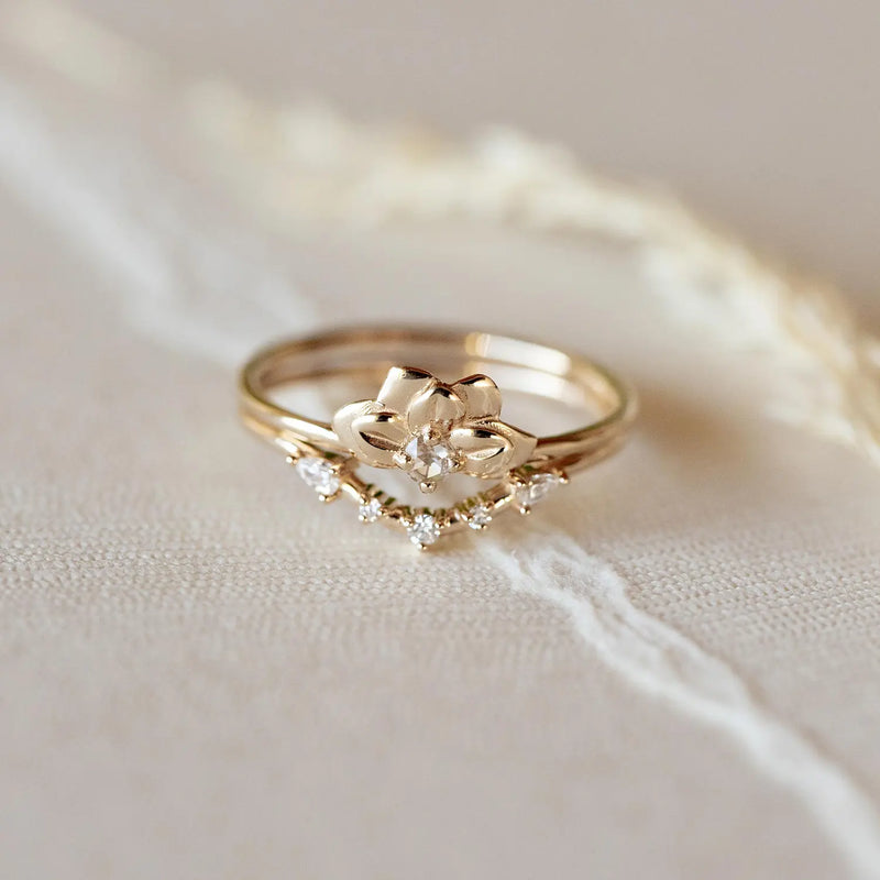 A close-up photo of the "A deep love band" from beloved by bluboho, an elegant 14k yellow gold ring, set on a soft beige surface. This focal ring features a delicate floral design with a central white diamond and several smaller white diamonds around it, casting a subtle sparkle.