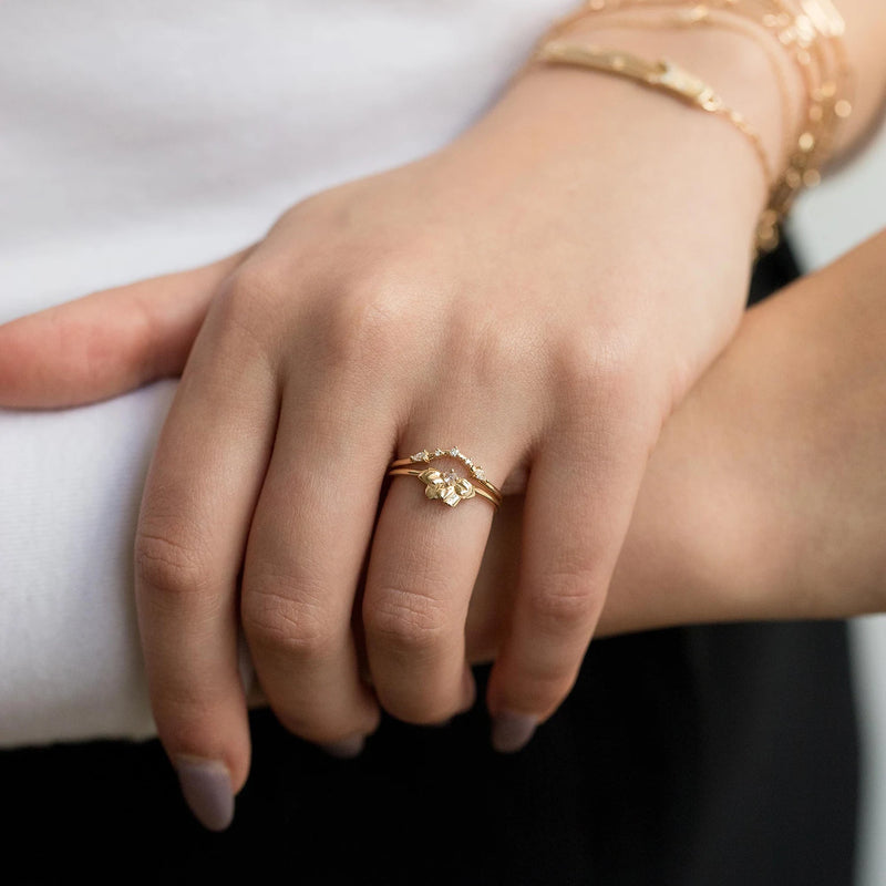 A close-up of a hand wearing "A deep love band" by beloved by bluboho, an elegant 14k yellow gold ring featuring white diamonds in a delicate flower design. The person is also wearing a white shirt and gold bracelets on their wrist.