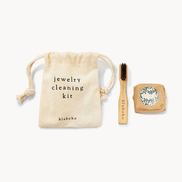 A sustainable non-toxic cleaning kit from bluboho, featuring a small drawstring bag labeled "sustainable non toxic cleaning kit" and "bluboho," a wooden brush with dark bristles, and a bar of soap wrapped in paper with a decorative label. The items are arranged neatly on a white background.