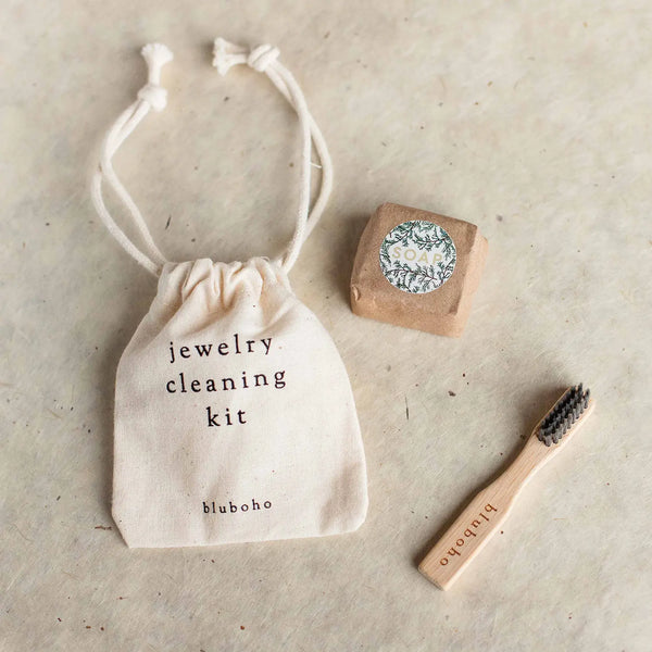 A beige drawstring bag labeled "sustainable non toxic cleaning kit" lies on a light surface along with a small bar of soap wrapped in paper and a wooden cleaning brush with black bristles. The "bluboho" branding is visible on both the brush and the bag.