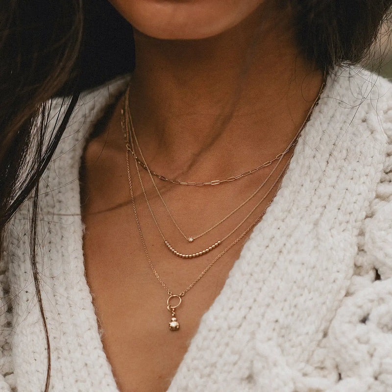 Close-up of a person wearing several layered necklaces, including the Dainty Diamond Nova Necklace by bluboho in 14k yellow gold. The necklaces vary in length and feature different pendants, such as a small gold charm and a delicate circular pendant. The person is dressed in a white, textured garment.