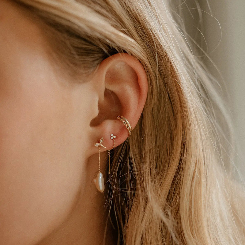 Close-up of a woman's ear adorned with multiple earrings, including the elegant bluboho abacus tripod diamond earring in 14k yellow gold with a white diamond. She is also wearing a hoop, a small stud with a floral design, and a drop earring featuring a pearl. Her light-blonde hair cascades around her ear while the background remains softly blurred.