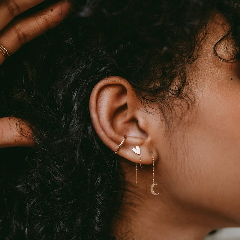 Close-up of a person's ear adorned with multiple earrings. The earrings include a small hoop, the Everyday Larger Lovely Heart Earring in 14k yellow gold by bluboho, a dangling bar, and a delicate chain with a crescent moon charm. The person's hand is gently touching their curly hair.