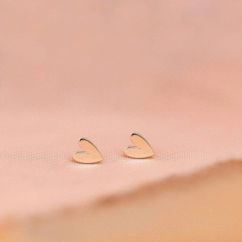 The Everyday Larger Lovely Heart Earrings from bluboho, crafted in elegant 14k yellow gold, are set against a soft pink background. These earrings feature a simple, minimalist design that exudes refined elegance.