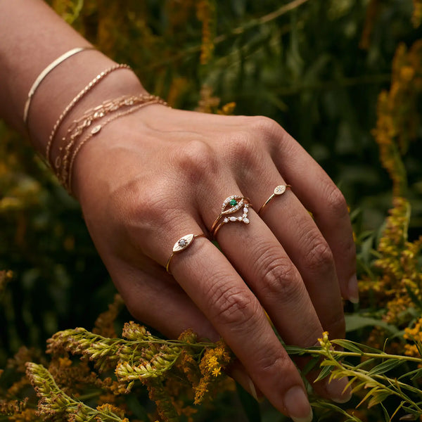 A hand adorned with multiple rings featuring a mix of gemstones and designs, including the stunning visionary evil eye ring - 14k yellow gold with brilliant diamond from bluboho, and delicate chains on the wrist. The background is filled with lush, yellow-green foliage, adding a natural element to the composition.