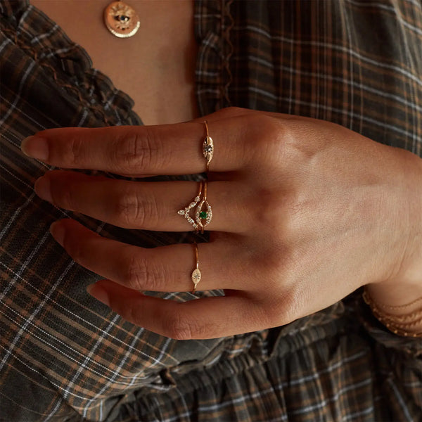 A close-up of a person's hand adorned with the bluboho tsavorite mystical evil eye ring, featuring the vibrant green tsavorite garnet set in 14k yellow gold. The person is wearing a plaid blouse with ruffled trim and a gold pendant necklace. The backdrop is softly lit, highlighting the jewelry and hand.