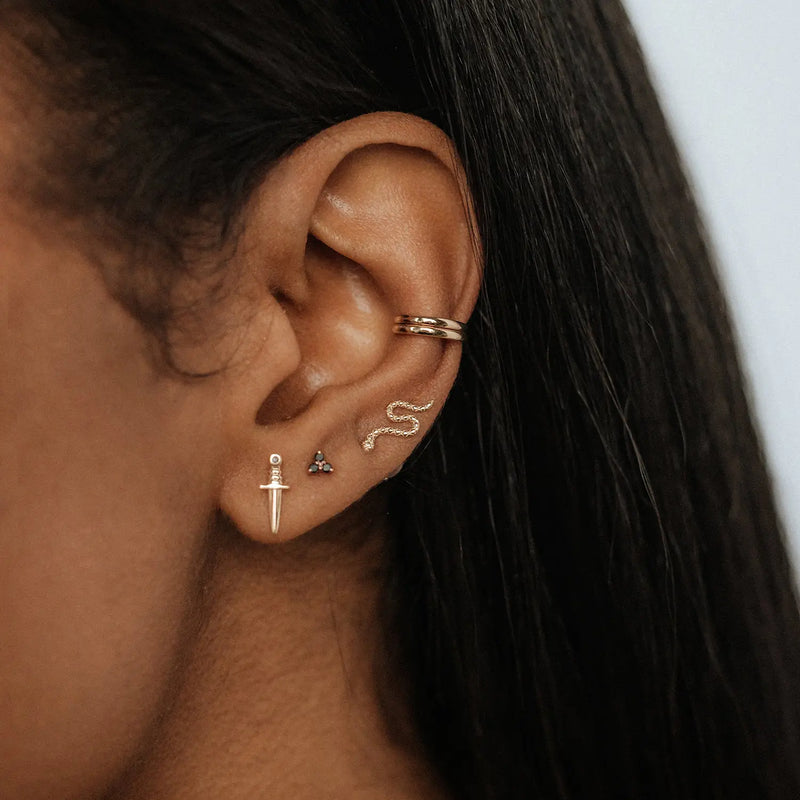 Close-up of a person's ear adorned with multiple earrings, including a small snake-shaped earring, a tiny jewel stud, a dagger-shaped earring, two small hoop earrings at the top of the ear, and the Guardian ear cuff from bluboho in 14k yellow gold. The person has dark hair.