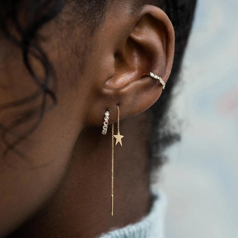 A close-up of a person's ear adorned with multiple gold earrings from bluboho. The ear lobe features a hoop earring with small gems, a Guidance Star Threader in 14k yellow gold, and an upper ear cuff also embellished with small gems. The person has dark skin and curly hair that is partially visible.