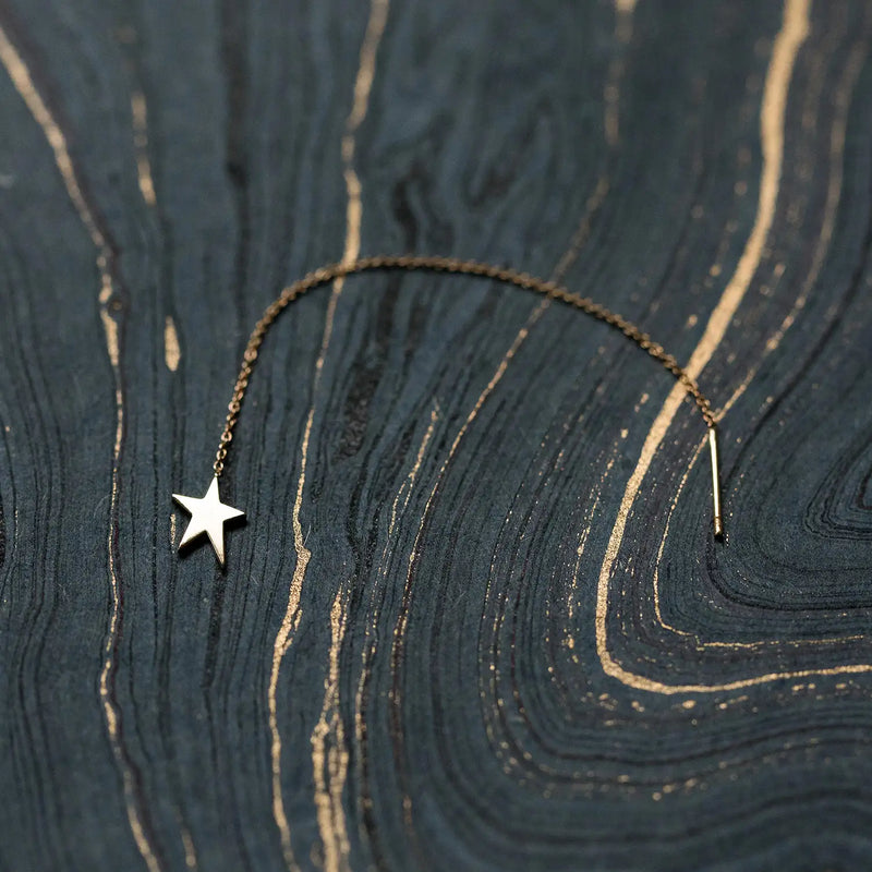 The Guidance Star Threader by bluboho, crafted from 14k yellow gold, features a delicate chain with a small, shiny star-shaped charm at one end and a thin bar at the other. It is elegantly displayed on a dark, textured surface with swirling patterns.