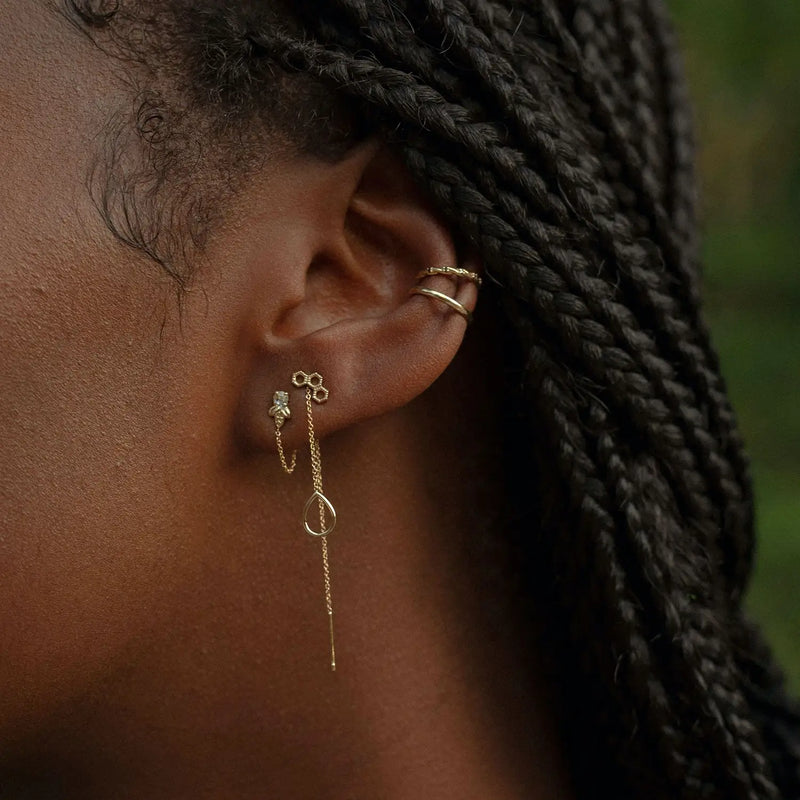 Close-up of a person's ear adorned with multiple bluboho earrings. The ear has a gold hoop in the cartilage, small gold studs, and a long, thin gold chain earring with a Honeycomb earring - 14k yellow gold teardrop-shaped accent in the lobe. The person has braided hair.