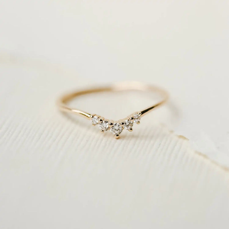 The Kismet Serendipity ring by bluboho, crafted in 14k yellow gold and featuring white diamonds in a V-shaped design, is displayed on a textured white surface. The simplicity and elegance of this ring make it a graceful piece of jewelry.