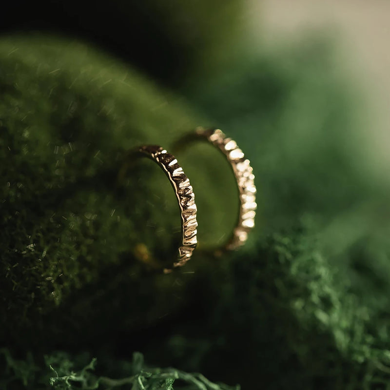 A pair of Larger Tree Bark Stability Hoops in 14k yellow gold from bluboho sit closely together on a lush green surface. The intricate detailing on the rings contrasts with the smooth, rich fabric beneath, creating an elegant and luxurious feel.