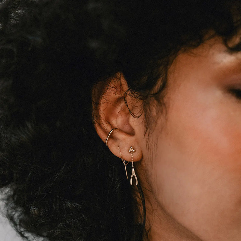 A close-up side profile of a person's ear adorned with two earrings. The upper earlobe has a gold hoop, while the lower lobe features the Large Abacus Tripod Earring in 14k yellow gold from bluboho, showcasing a delicate wishbone shape with a small attached gemstone near the piercing. The person has curly dark hair.