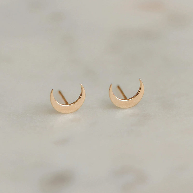 Close-up of a pair of Everyday larger crescent moon earrings by bluboho, crafted in 14k yellow gold, on a light speckled surface. The earrings have a simple, polished finish giving them a sleek and elegant appearance.