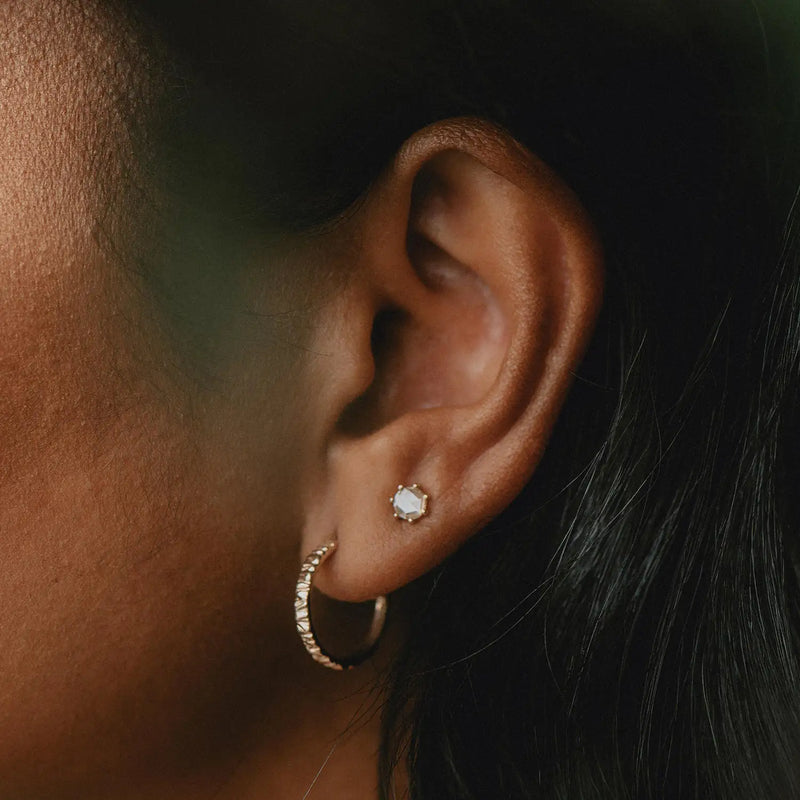 Close-up of a person's ear adorned with two earrings: a small, round stud with a gem at the top and the bluboho Larger Tree Bark Stability Hoop in 14k yellow gold below. The background is blurred, focusing attention on the jewelry.