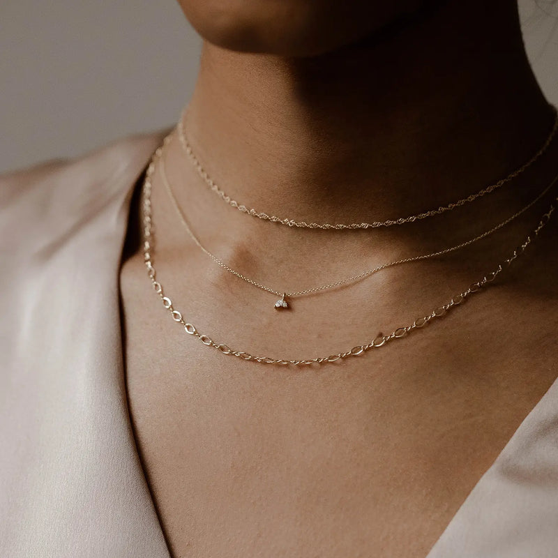 Close-up of a person wearing three delicate, layered gold necklaces. One necklace, identified as the Lean on me diamond necklace in 14k yellow gold by bluboho, features a small triangular pendant. The other two are simple chains with varied links. The person is dressed in a silky, light-colored blouse.