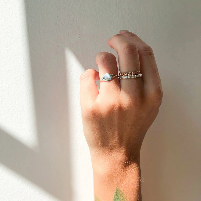 A hand adorned with three rings is held up against a white wall, bathed in natural sunlight that casts gentle shadows. Among the rings, one showcases a stunning blue stone while the other two feature sleek band designs. Also visible on the wrist is a green tattoo. The elegant bands include the Loyalty signet ring by bluboho, crafted from sterling silver and engravable for a personal touch.