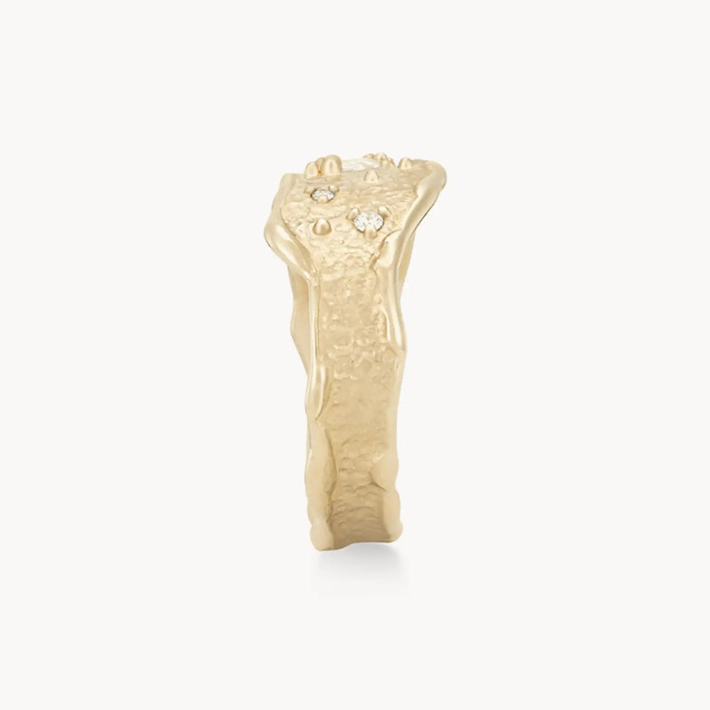 The Luminous Honey Diamond Ring by bluboho, crafted from 14k yellow gold, features a rough, natural finish and an organic, uneven shape. Three small white diamonds are embedded on the surface, adding to its unique and handcrafted appearance. The ring is set against a plain white background.