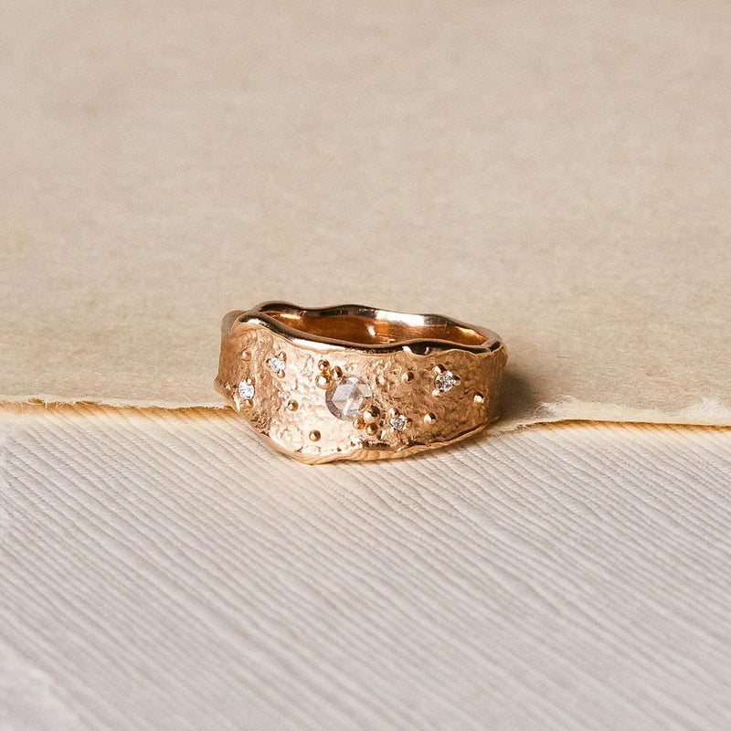 The Luminous honey diamond ring by bluboho, crafted from 14k yellow gold and adorned with white diamonds, rests on a beige background. The ring features an uneven, textured surface that exudes rustic charm, while the embedded gemstones add elegance and shine. A subtle crease in the background enhances the minimalistic and neutral setting.