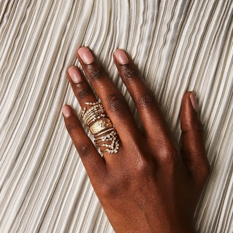 A close-up of a person's hand with dark skin tone, resting on a textured white surface. The hand is adorned with several gold rings featuring various delicate designs and intricate details, including the Luminous Honey Diamond Ring by bluboho in 14k yellow gold with white diamonds. The nails are manicured with a light pink polish.