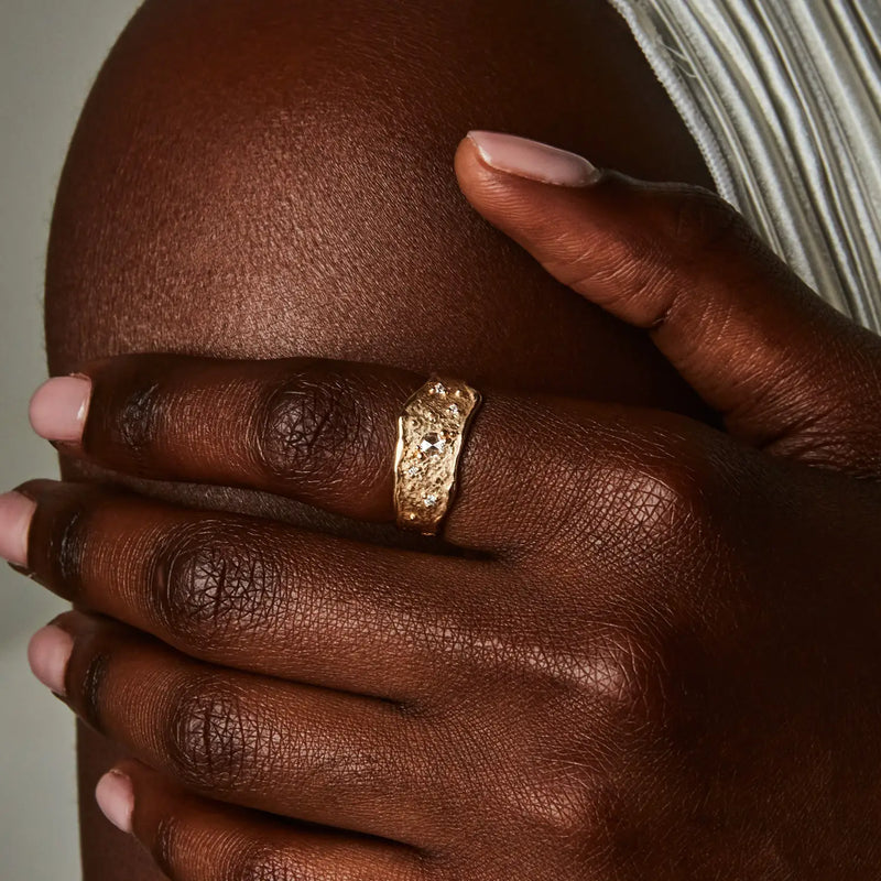 A close-up of a hand with dark skin wearing the Luminous Honey Diamond Ring by bluboho, crafted in 14k yellow gold and adorned with white diamonds. The fingers are touching an upper arm that is also visible in the image. The person's nails are manicured with light pink polish, and a white garment is partially shown.