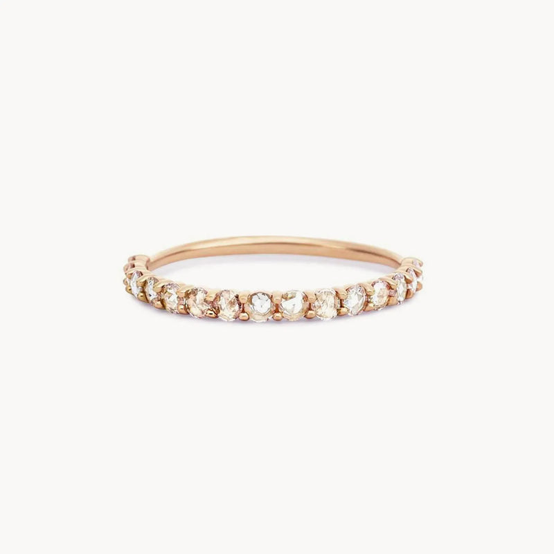 The Moonglade ring by beloved by bluboho is a delicate 14k rose gold piece, adorned with small, round white diamonds set in a row across the top. The thin and elegant band enhances the sparkle of the diamonds, while a white background highlights its intricate design.