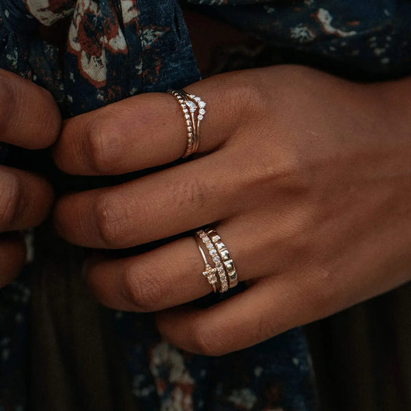 Close-up of two hands adorned with various Moonglade rings from beloved by bluboho, crafted in 14k rose gold and set with white diamonds. The person's unpolished nails are visible against the dark, floral-patterned fabric in the background. The rings are elegantly stacked in different styles on multiple fingers.