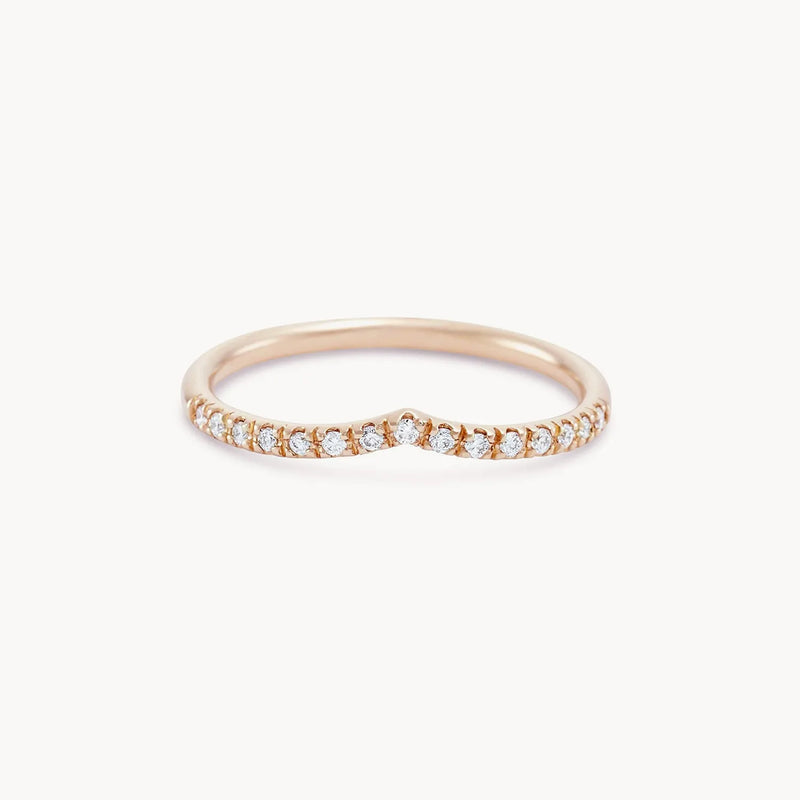 The Peak Eternity Band by Beloved by Bluboho is a delicate 14k rose gold ring featuring a subtle V-shaped curve at the center. Adorned with small, sparkling white diamonds set along the top half of the band, it boasts an elegant and graceful design against a plain white background.