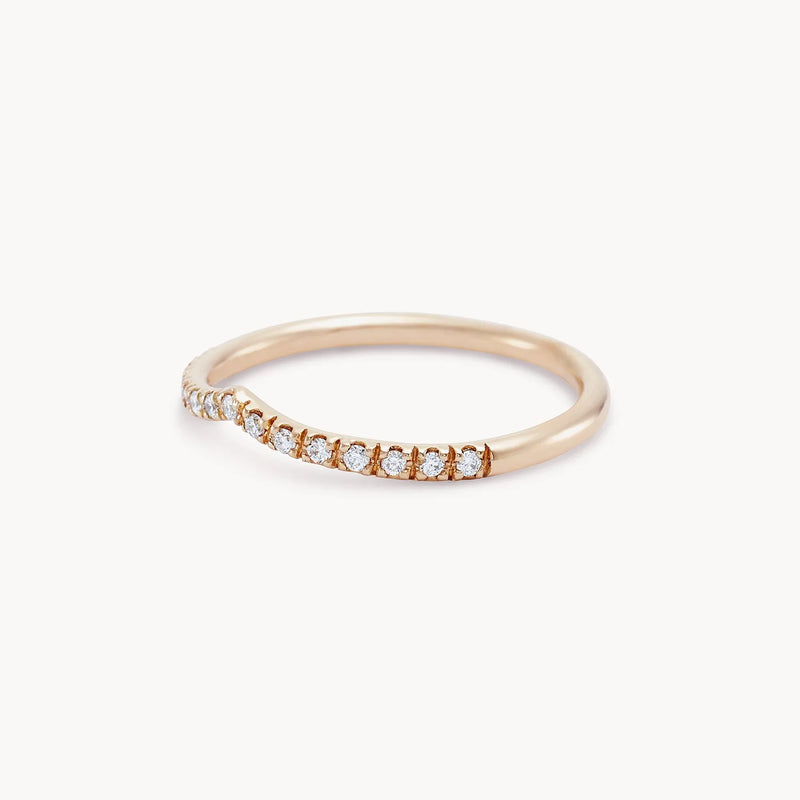 The peak eternity band by beloved by bluboho is a delicate 14k rose gold ring featuring a subtle curve adorned with a row of small, sparkling white diamonds along the top half. It has a thin and elegant band, offering a minimalist yet sophisticated design against a plain white background.