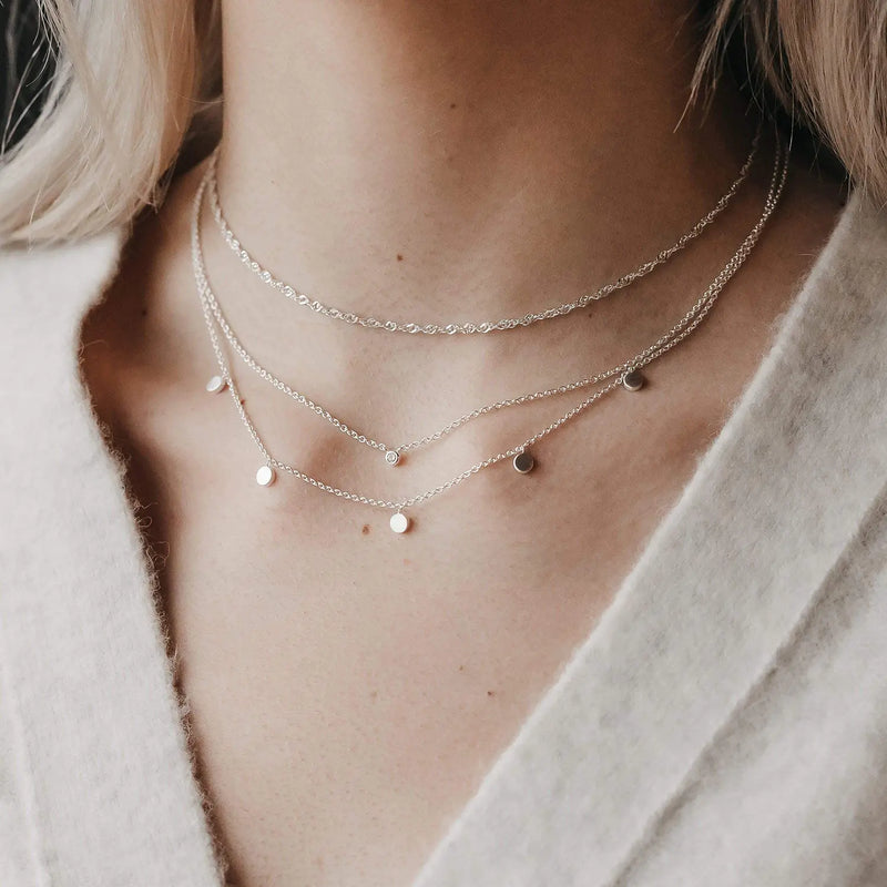 A close-up image showing a person adorned with the bluboho lightning strikes choker necklace in sterling silver, styled as part of a trio of layered necklaces. The first is a delicate chain, the second features small round discs, and the third is a simple chain with small, spaced discs. The person is dressed in textured light-colored fabric.