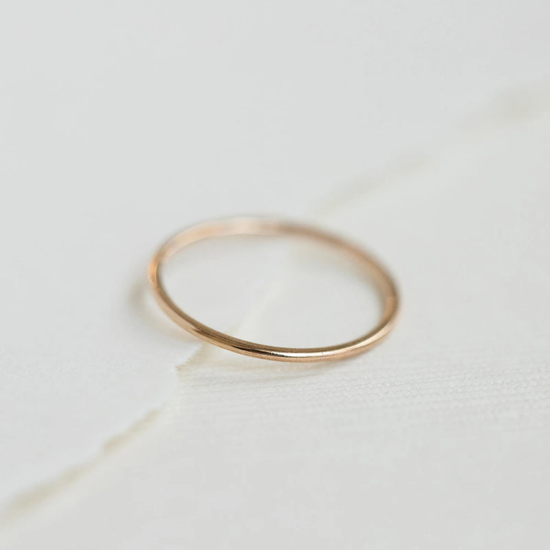 A plain jane stacking ring in 10k yellow gold from bluboho is placed on a white, textured surface. The ring showcases a minimalistic design with a simple, smooth band, free of any additional decorations. The softly lit background emphasizes the delicate nature of the ring.