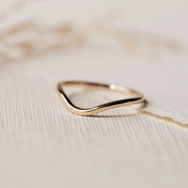 A plain wave band - 14k yellow gold by beloved by bluboho lies on a textured white surface. The background is softly blurred, drawing attention to the delicate and elegant shape of the ring.