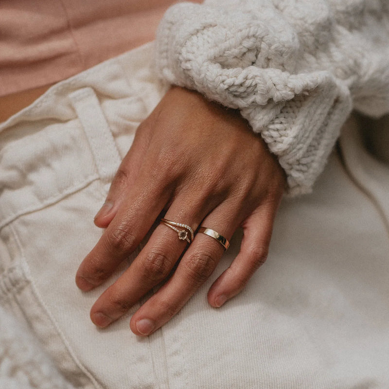 A close-up of a hand resting on the edge of white jeans. The hand wears two rings: one is a delicate, thin, twisted band, and the other is the wave eternity ring in 14k yellow gold with white diamonds by beloved by bluboho. The person is wearing a white chunky knitted sweater.