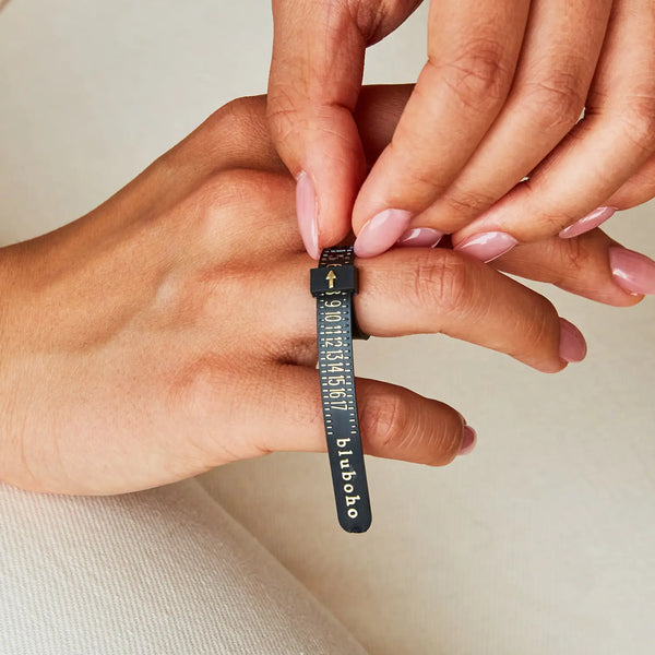 Close-up of two hands, one using a black plastic Bluboho ring sizer to measure the ring size of a finger. The hand holding the sizer has a pale manicure. The sizer has white numbers and markings, with the text "bluboho" visible. The background is neutral.