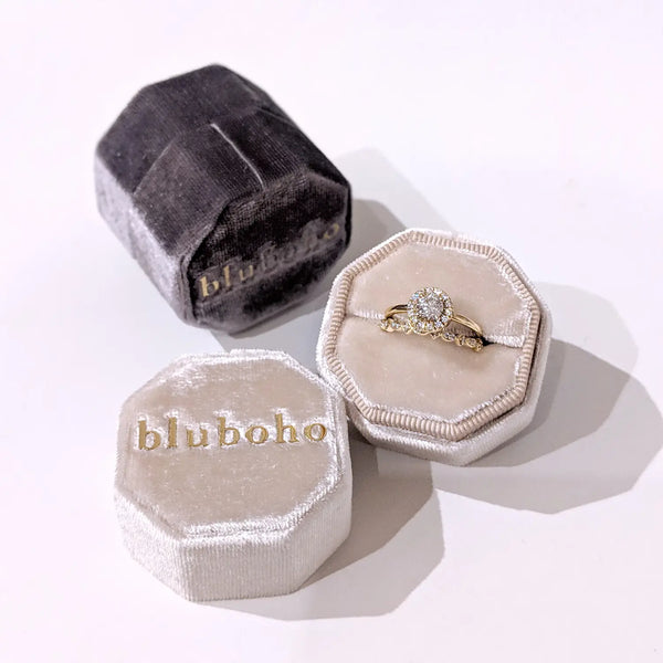 A delicate gold ring adorned with a round diamond center stone and smaller diamonds on the band is nestled in a plush, hexagonal Beloved by bluboho Box - Velvet. The grayish velvet ring box, branded with "beloved by bluboho" in gold lettering, has its lid removed and placed beside it.