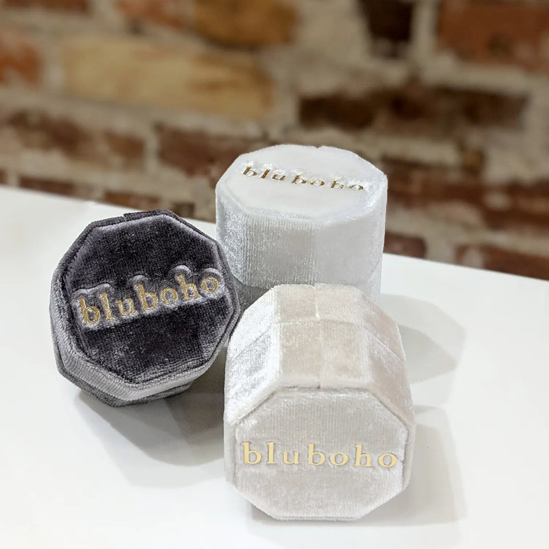 Three octagonal Beloved by bluboho Velvet jewelry boxes in gray, off-white, and cream with "bluboho" printed in gold lettering on the lids are displayed on a white surface with a brick wall in the background.