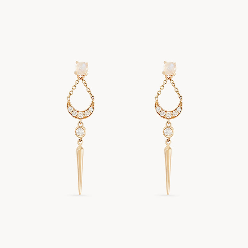 The sacred geometry moonstone earring by bluboho is a pair of elegant, 14k yellow gold earrings featuring a small round stud connected to a crescent-shaped design adorned with sparkling crystals. From the crescent, a chain with a larger crystal and a pointed gold spike dangles delicately against the white background.