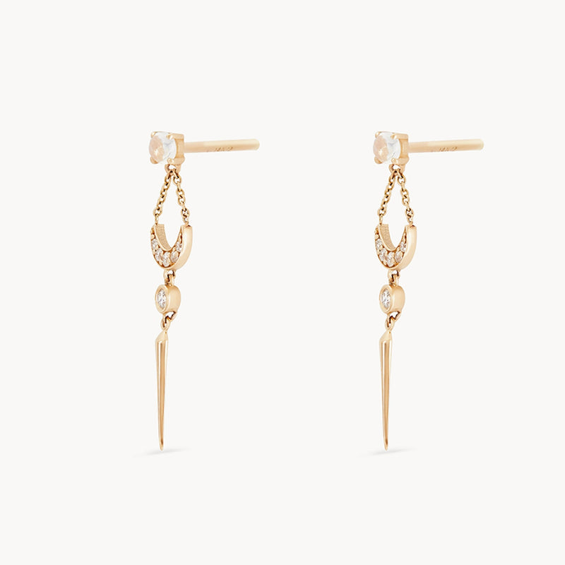 The sacred geometry moonstone earrings by bluboho are a pair of elegant 14k yellow gold earrings featuring a double-drop design. Each earring has a small stud with a curved element adorned with tiny moonstones, connected to a pointed spike hanging at the bottom, all set against a plain white background.