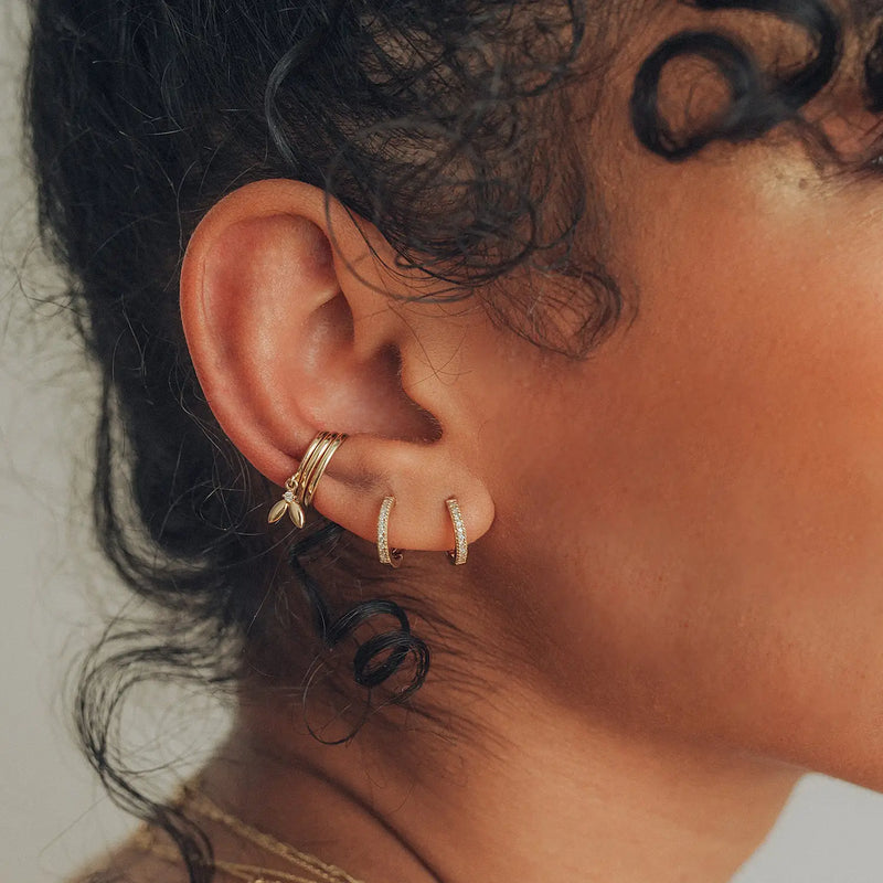 Close-up of a person’s ear adorned with jewelry. The ear has several piercings, featuring three small gold hoop earrings on the upper cartilage and two larger bluboho shine on hoop earrings - 14k yellow gold, white diamond with embedded gems on the earlobe. The person has curly hair.