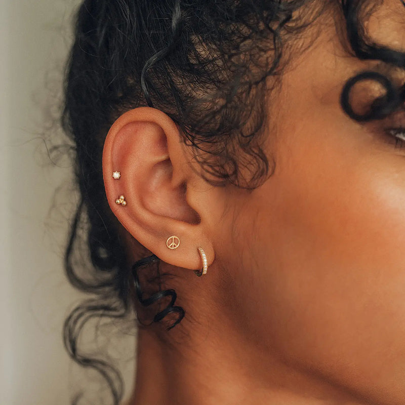 Close-up of a person's ear adorned with multiple earrings. The ear features three piercings: two small stud earrings and one bluboho shine on hoop earring - 14k yellow gold, white diamond with a peace symbol charm. The person has dark, curly hair partially visible around the ear.