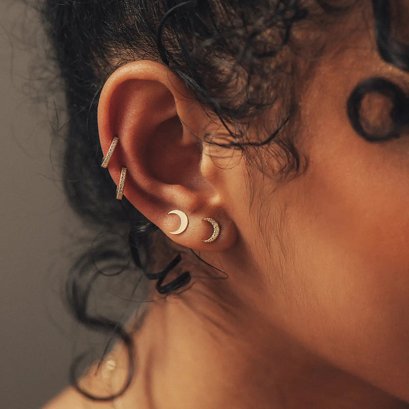 Close-up of a person's ear adorned with multiple earrings. The earrings include a pair of Bluboho Shine On Hoop Earrings - 14k Yellow Gold, White Diamond on the earlobe and two small bar-shaped studs in the cartilage. The person's black curly hair partially frames the ear.