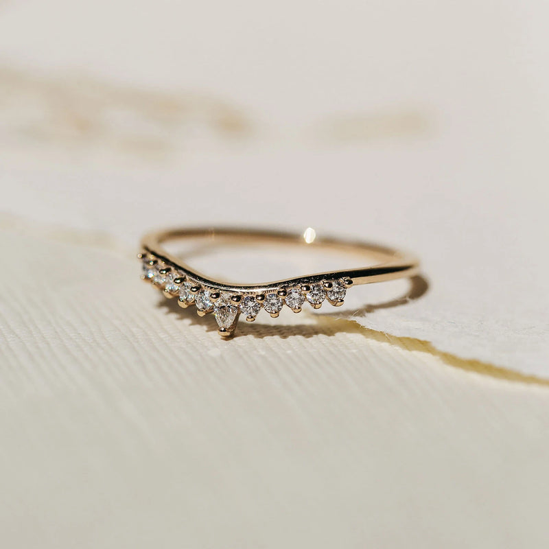 A delicate sunbeam ring in 14k yellow gold with a V-shaped design, adorned with small, shimmering white diamonds along its top edge by beloved by bluboho, placed on a textured, light-colored surface.