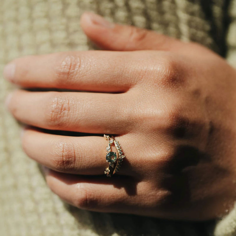 A hand adorned with the Sunbeam Ring by Beloved by Bluboho—a stunning piece crafted from 14k yellow gold, featuring a green gemstone and small white diamonds—rests on a textured surface. The sunlight illuminates the intricate design of the ring. The skin appears smooth, and the fingers are elegantly positioned to showcase the jewelry beautifully.