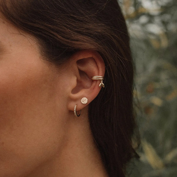 Close-up of a person's ear adorned with multiple gold earrings. The ear features a small hoop in the earlobe, a Tree of Life Stud Earring - 14K Yellow Gold by bluboho above the lobe, and two additional small hoops with dangling charms on the cartilage. The background is blurred foliage.