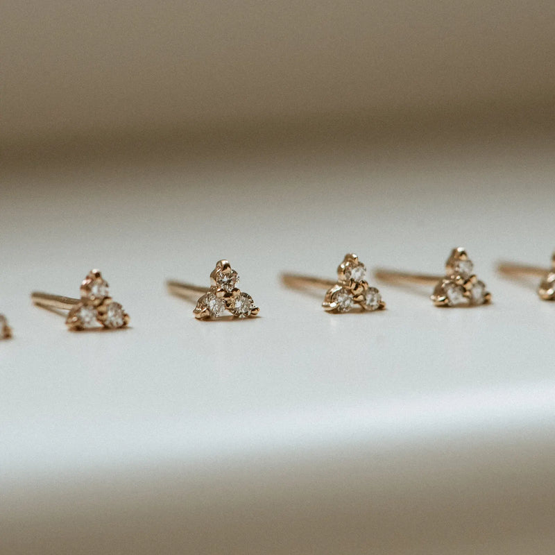 Several abacus tripod diamond earrings by bluboho, featuring clusters of three small white diamonds set in 14k yellow gold and forming a triangular shape, are arranged in a neat row on a smooth, neutral-colored surface. The earrings are displayed under soft, diffused lighting, highlighting the sparkle of the diamonds.