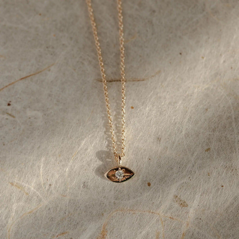 Close-up of the visionary evil eye pendant - 14k yellow gold by bluboho, featuring a small gemstone at its center. The delicate gold necklace is displayed on a textured, light-colored surface.