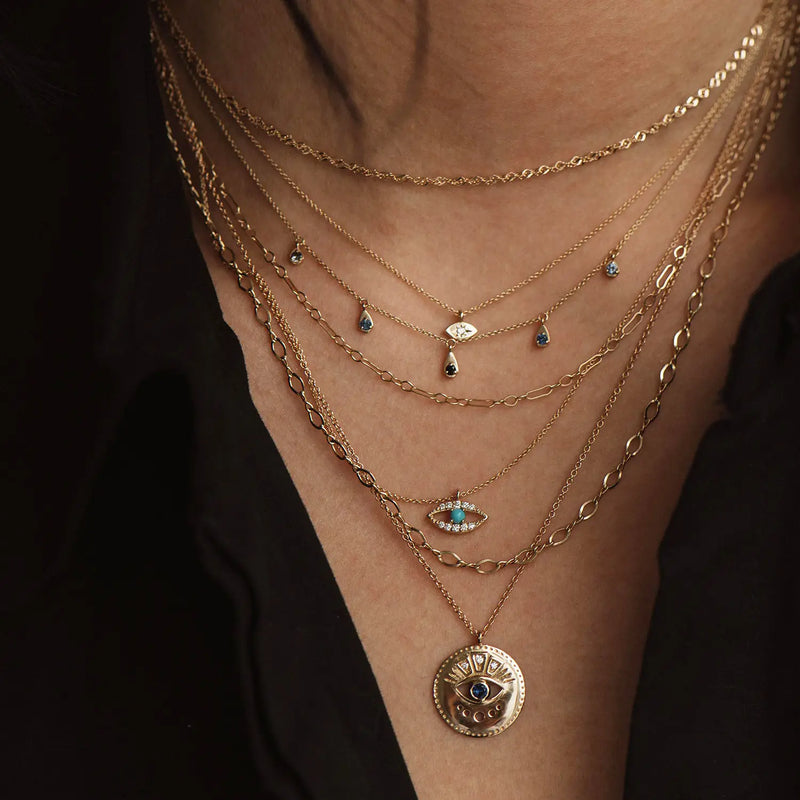 Close-up of a person's neck adorned with several layered gold necklaces, including the visionary evil eye pendant by bluboho. These 14k yellow gold necklaces showcase various delicate designs such as a finely detailed gold disc pendant, an evil eye charm, small teardrop charms, and fine chain links, creating an elegant appearance.