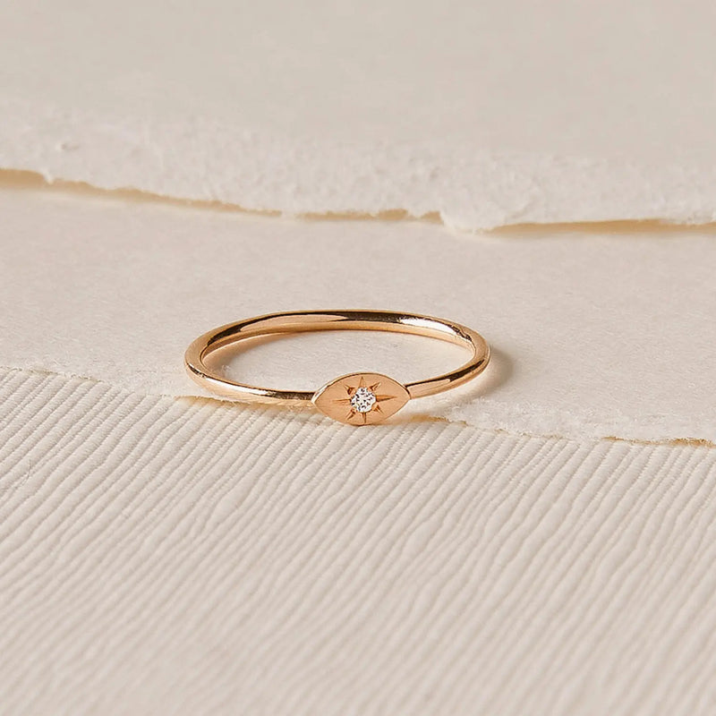 A delicate visionary evil eye ring in 14k yellow gold lies on a textured cream-colored background. The ring, by bluboho, features an oval-shaped centerpiece with a small, brilliant diamond set in the center. The backdrop includes layers of paper with varying textures.